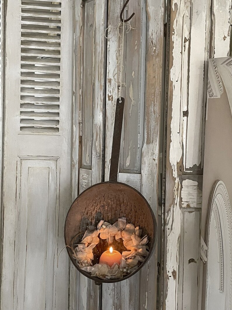 May include: A rustic metal colander hanging from a hook, with a white lace wreath and a lit candle inside. The colander is hanging against a whitewashed wooden wall.