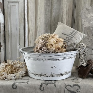 May include: A white metal basin with distressed paint and the number 45 printed on the front. The basin features two side handles and is filled with a cluster of dried beige flowers, accompanied by a small rectangular fabric pillow.