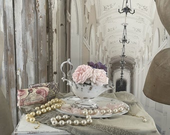 May include: A white ceramic bowl with a decorative handle, filled with pink and purple flowers. The bowl is sitting on a white table with a pearl necklace and a plate with pink flower petals. The background is a white hallway with a chandelier.