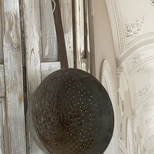 May include: A vintage metal colander with a long handle hanging on a white wooden wall. The colander has a distressed finish and is covered in small holes.