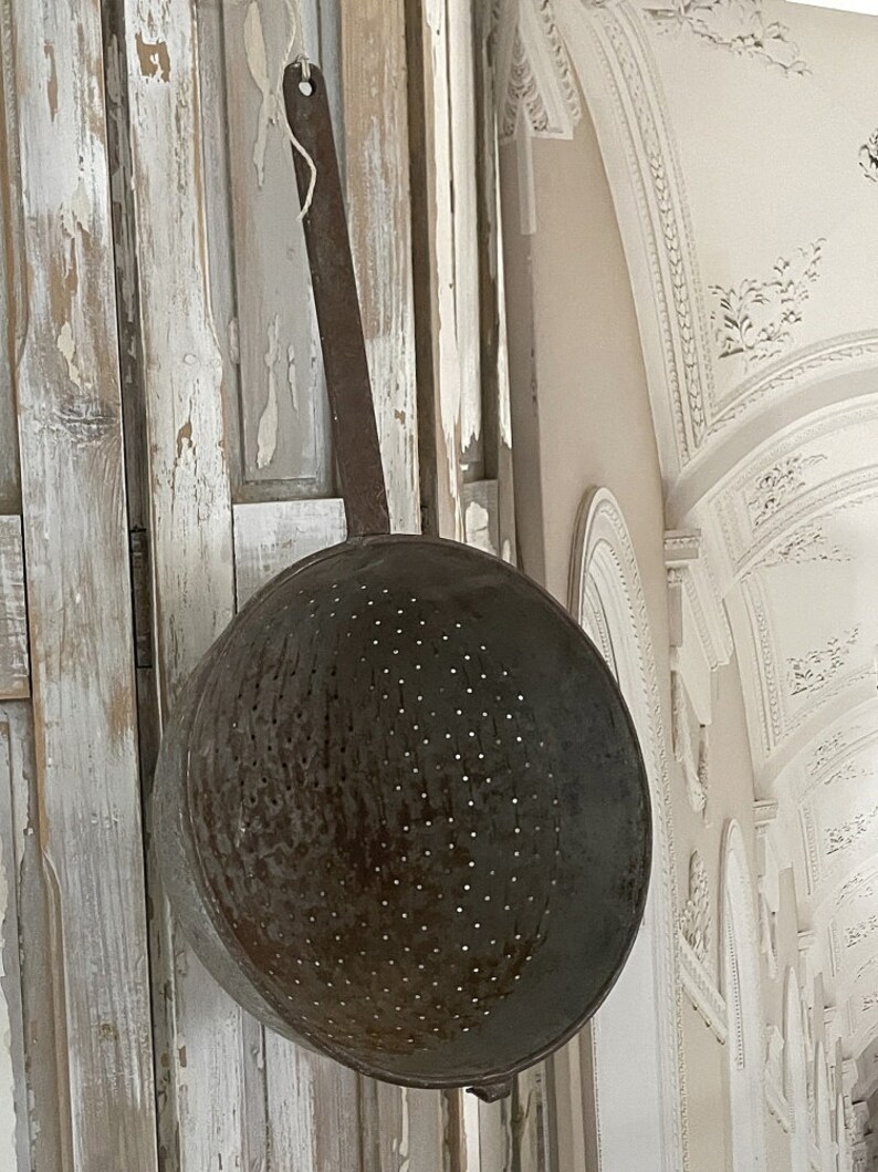 May include: A vintage metal colander with a long handle hanging on a white wooden wall. The colander has a distressed finish and is covered in small holes.