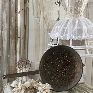 May include: A vintage colander with a long handle sits on a white fabric surface with a white lace trim. A white fabric wreath with a shell design is in the foreground. A white dress form with a white lace dress is in the background.