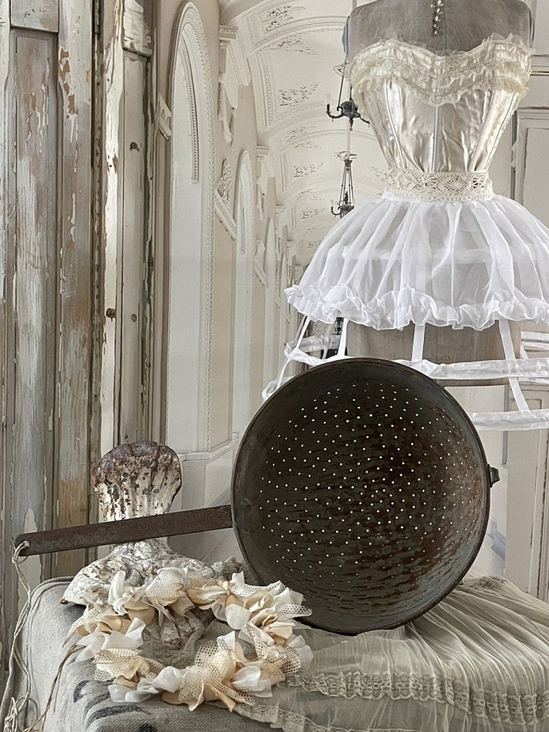 May include: A vintage colander with a long handle sits on a white fabric surface with a white lace trim. A white fabric wreath with a shell design is in the foreground. A white dress form with a white lace dress is in the background.