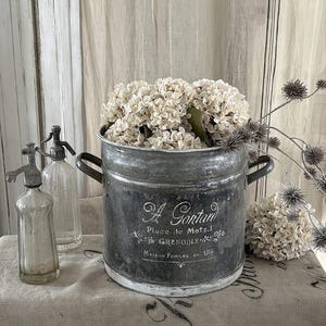 Puede incluir: Un cubo de metal desgastado con asas, lleno de flores de color crema. El cubo tiene texto en francés y está junto a dos botellas de sifón de vidrio vintage. La escena está ambientada sobre una mesa cubierta de lino.
