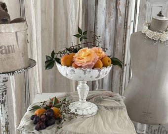 May include: A white, distressed pedestal fruit bowl filled with peaches and peach-colored roses. The bowl sits on a table with a draped cloth, next to a dress form and a vintage hat box. The scene has a rustic, vintage aesthetic.