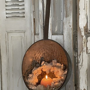 May include: A vintage metal colander with a rustic finish, hanging from a metal handle. The colander is filled with a delicate white lace wreath and a single orange candle.