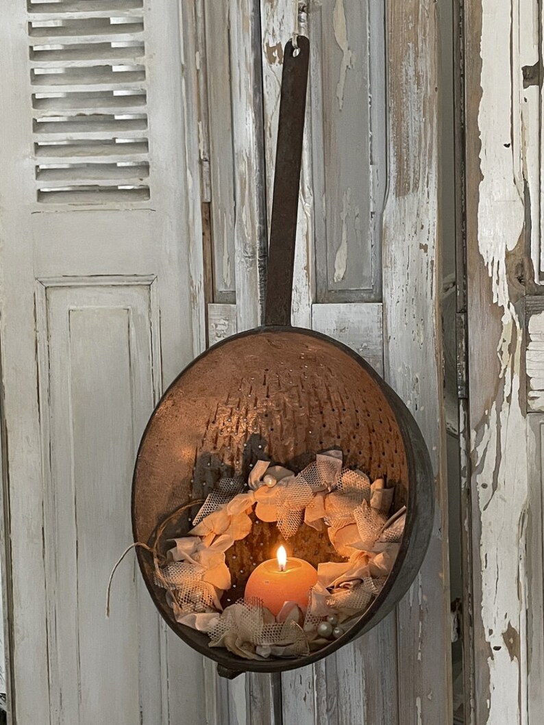 May include: A vintage metal colander with a rustic finish, hanging from a metal handle. The colander is filled with a delicate white lace wreath and a single orange candle.