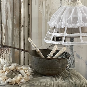 May include: A white ruffled lampshade with a cage-like base sits on a table with a rustic metal bowl filled with rolled up sheet music. A white lace wreath sits on the table in front of the bowl.