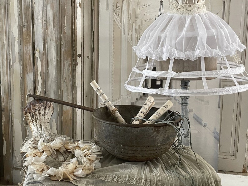 May include: A white ruffled lampshade with a cage-like base sits on a table with a rustic metal bowl filled with rolled up sheet music. A white lace wreath sits on the table in front of the bowl.