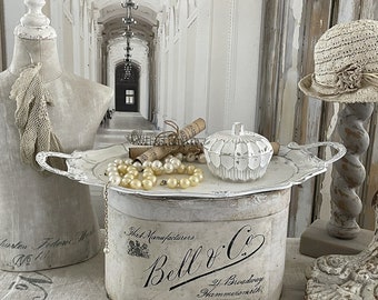 May include: A white ceramic trinket box with a fluted design sits on a white tray with silver handles. The tray is on top of a white box with black lettering that reads "Hat Manufacturers Bell & Co. 21 Broadway Hammersmith". The box is surrounded by a string of pearls and a white lace scarf.