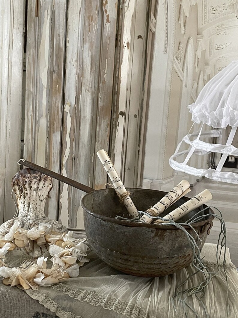 May include: A vintage metal colander with a rustic finish, filled with rolled sheet music and tied with twine. The colander is sitting on a white lace tablecloth with a white, lace-trimmed, decorative object in the foreground.
