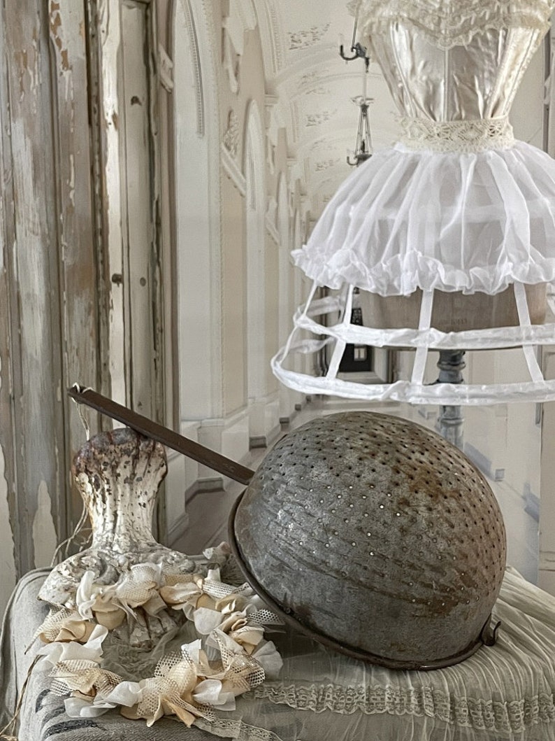 May include: A vintage metal colander with a long handle, sitting on a white lace tablecloth with a white lace crinoline in the background.
