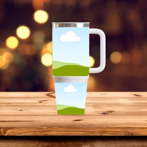 May include: A stainless steel travel mug with a white handle. The mug features a landscape design with a blue sky, white clouds, and green rolling hills. The mug is sitting on a wooden surface.