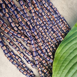 Könnte beinhalten: Eine Strähne aus blauen, weißen, orangefarbenen und gelben, gestreiften Glasperlen. Die Perlen sind in einer Reihe angeordnet und alle gleich groß.