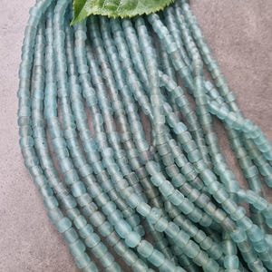 Könnte beinhalten: Stränge aus hellblauen, zylindrischen Perlen. Die Perlen sind aufgereiht und bilden mehrere Reihen. Ein grünes Blatt liegt auf den Perlen. Die Perlen haben ein leicht durchscheinendes Aussehen.