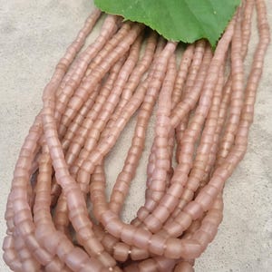 Könnte beinhalten: Mehrere Stränge aus kleinen, matten, altrosa farbenen Perlen, die in einem langen, fließenden Bündel angeordnet sind. Die Perlen haben eine weiche, halbtransparente Oberfläche und eine gleichmäßige, leicht abgerundete zylindrische Form, gekrönt von einem großen grünen Blatt.