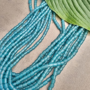 May include: A strand of turquoise blue glass beads, each bead is a cylinder shape with rounded edges. The beads are arranged in a loose pile, creating a flowing, organic look.