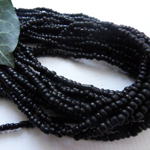 Peut inclure: Gros plan sur des brins de petites perles noires mates. Les perles sont enfilées ensemble, formant plusieurs rangées. Une feuille verte est visible dans le coin supérieur gauche, ajoutant un élément naturel à la composition. Les perles sont probablement utilisées pour la fabrication de bijoux.