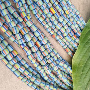 Op de afbeelding: Een close-up van een streng gekleurde glazen kralen. De kralen zijn klein en cilindrisch, met afwisselende strepen in blauw, geel en rood. De kralen zijn in een willekeurig patroon gerangschikt, wat een visueel aantrekkelijk ontwerp creëert.