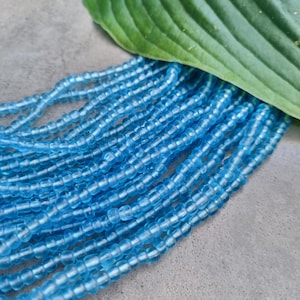 Könnte beinhalten: Nahaufnahme einer Strähne aus hellblauen Glasperlen. Die Perlen sind klein und rund und zu einer langen, fließenden Strähne angeordnet.