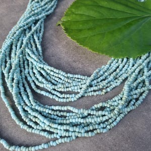Könnte beinhalten: Eine mehrsträngige Halskette, die aus zahlreichen kleinen, hellblauen Saatperlen in einem kaskadierenden, geschichteten Design besteht. Die Perlen haben eine matte Oberfläche und sind so aufgereiht, dass sie ein voluminöses, strukturiertes Erscheinungsbild erzeugen.