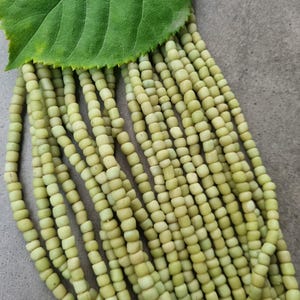 Könnte beinhalten: Stränge hellgrüner, zylindrischer Perlen, jeweils etwa 0,5 cm lang, liegen unter einem großen grünen Blatt. Die Perlen haben eine leicht unregelmäßige Form und ein mattes Finish. Die Anordnung deutet auf ein Bastel- oder Schmuckthema hin.