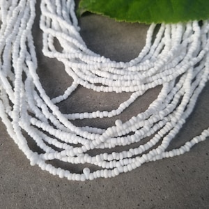 Könnte beinhalten: Nahaufnahme von mehreren Strängen weißer Rocailles-Halsketten. Die kleinen, gleichmäßigen Perlen erzeugen ein zartes, strukturiertes Aussehen. Ein großes grünes Blatt liegt im Hintergrund und setzt einen natürlichen Kontrast zu den hellweißen Perlen.