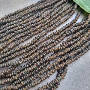 Op de afbeelding: Een streng donkergroene en bruine glazen kralen. De kralen zijn klein en rond en zijn aan elkaar geregen op een doorzichtige draad.