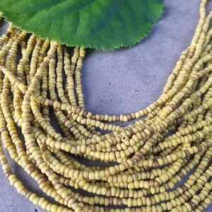 Könnte beinhalten: Nahaufnahme einer mehrsträngigen Perlenkette. Die Perlen sind hauptsächlich gelb mit einigen braunen Perlen dazwischen. Im Hintergrund liegt ein großes grünes Blatt auf einer grauen Oberfläche. Die Halskette ist wahrscheinlich handgefertigt.