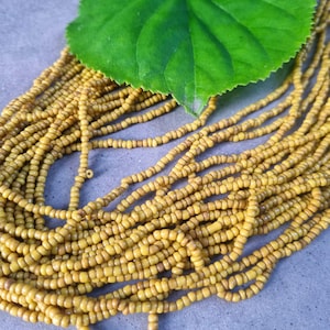 May include: A close-up shot of multiple strands of yellow beads, likely for jewellery. A large, vibrant green leaf sits atop the beads. The beads are small and consistent in size, with a slightly matte finish. The background is a muted grey.