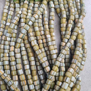 Könnte beinhalten: Nahaufnahme von mehreren Strängen zylindrischer Perlen. Die Perlen sind gelb mit Streifen in Weiß, Orange, Grün und Schwarz. Oben liegt ein grünes Blatt. Ideal für die Schmuckherstellung oder Bastelprojekte.