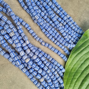 Könnte beinhalten: Nahaufnahme einer Schnur aus blauen und weißen, gestreiften Glasperlen. Die Perlen sind fächerförmig angeordnet und liegen auf einer grauen Oberfläche. Ein grünes Blatt ist im Hintergrund teilweise sichtbar.