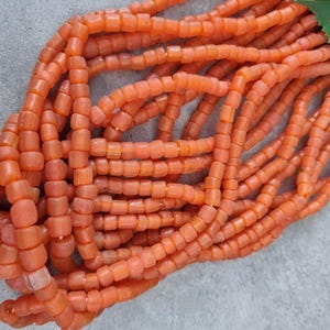 Könnte beinhalten: Eine Ansammlung mehrerer Stränge aus kleinen, zylindrischen orangefarbenen Perlen. Die Perlen haben eine matte, leicht texturierte Oberfläche und sind in langen, parallelen Linien aufgereiht. Ein einzelnes grünes Blatt ist oben rechts zu sehen.