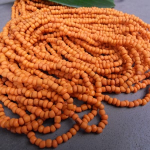 Könnte beinhalten: Nahaufnahme von einem Haufen orangefarbener Perlen, wahrscheinlich für die Schmuckherstellung. Die kleinen, zylindrischen Perlen sind aufgefädelt und erzeugen ein strukturiertes Aussehen. Ein grünes Blatt ist im oberen Teil des Bildes zu sehen.