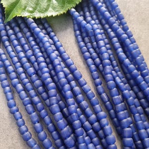 Könnte beinhalten: Nahaufnahme von mehreren Strängen blauer Glasperlen. Die Perlen sind klein, zylindrisch und haben eine matte Oberfläche. Ein großes grünes Blatt liegt auf den Perlen und bildet einen natürlichen Kontrast zum Blau.