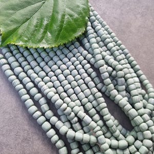 Könnte beinhalten: Mehrere Stränge hellgrüner Perlen mit Streifenmuster. Ein großes grünes Blatt liegt auf den Perlen. Die Perlen sind zylindrisch und scheinen aus Glas oder Keramik zu sein. Ideal für die Schmuckherstellung.