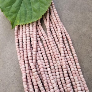 Könnte beinhalten: Stränge aus hellrosa quadratischen Perlen werden gezeigt, teilweise bedeckt von einem grünen Blatt. Die Perlen sind gleichmäßig in Größe und Form, wodurch ein texturierter Effekt entsteht. Der Hintergrund ist eine neutrale graue Oberfläche.