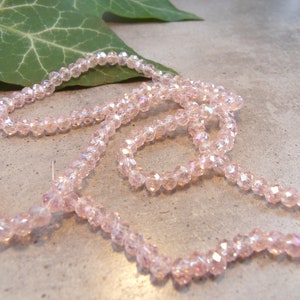 Könnte beinhalten: Stränge aus hellrosa, facettierten Perlen, vermutlich aus Kristall oder Glas, sind in einem fließenden Muster angeordnet. Die Perlen haben einen subtilen, irisierenden Schimmer. Ein grünes Blatt ist im Hintergrund zu sehen.