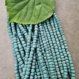 Könnte beinhalten: Nahaufnahme von Strängen türkisfarbener quadratischer Perlen. Ein großes grünes Blatt liegt auf den Perlen. Die Perlen sind aufgefädelt und bilden eine strukturierte und farbenfrohe Darstellung. Der Hintergrund ist ein neutrales Grau.