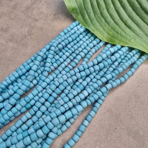 Könnte beinhalten: Eine Strähne aus hellblauen runden Perlen, die in Reihen angeordnet sind, vor einem grauen Hintergrund. Die Perlen sind klein und gleichmäßig groß.
