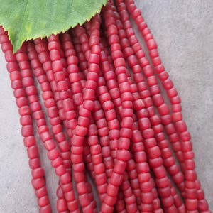 Könnte beinhalten: Stränge aus roten, quadratischen Perlen, möglicherweise aus Glas oder Stein, werden gezeigt. Ein grünes Blatt liegt oben und bedeckt die Perlen teilweise. Die Perlen haben eine tiefe, satte rote Farbe, und das Bild ist gut beleuchtet, wodurch die Textur und Form der Perlen hervorgehoben wird.