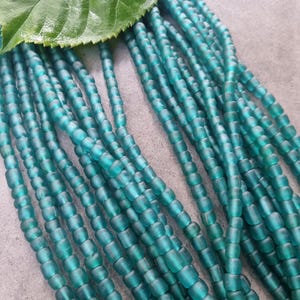 Könnte beinhalten: Nahaufnahme von türkisblauen Glasperlen mit mattem Finish, aufgefädelt. Die zylindrischen Perlen werden mit einem großen grünen Blatt präsentiert. Diese Perlen sind wahrscheinlich für die Schmuckherstellung bestimmt.