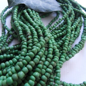 Può includere: Primo piano di più fili di perline verdi. Le perline sono piccole, rotonde e hanno una finitura opaca. Alcune perline presentano un aspetto più scuro e screziato. Una foglia verde è appoggiata sopra le perline. Le perline sono probabilmente di vetro o ceramica.