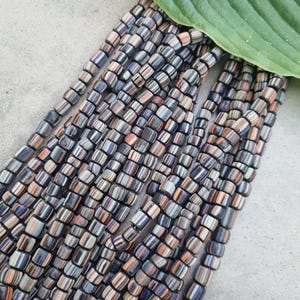 Op de afbeelding: Strengen rechthoekige kralen in tinten grijs, oranje en blauw. De kralen hebben een gestreept patroon en zijn in een trapsgewijze volgorde gerangschikt. Een groen blad is op de achtergrond.