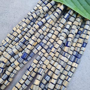 Pode incluir: Um fio de contas de vidro azul e creme. As contas são cilíndricas e têm um padrão listrado.