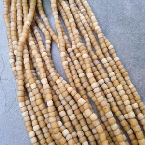 Könnte beinhalten: Nahaufnahme von mehreren Strängen hellbrauner, quadratischer Perlen. Die Perlen haben einen natürlichen, erdigen Ton mit Farbvariationen. Oben ist ein grünes Blatt zu sehen. Die Perlen sind wahrscheinlich für die Schmuckherstellung oder das Basteln gedacht.