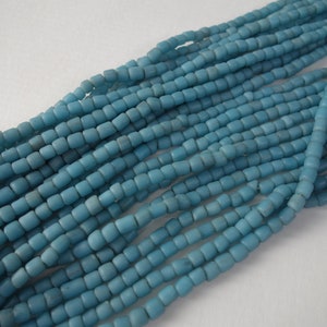 Pode incluir: Um fio de contas de vidro azul claro, cada conta tem formato quadrado com bordas arredondadas.