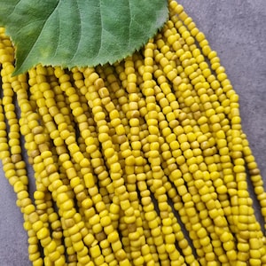 Könnte beinhalten: Nahaufnahme von Strängen gelber Perlen mit einem grünen Blatt. Die kleinen, zylindrischen Perlen haben ein mattes Finish. Die Perlen sind in einem kaskadierenden Muster angeordnet, wodurch ein texturierter Effekt entsteht.