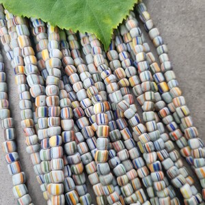 Könnte beinhalten: Nahaufnahme von Strängen zylindrischer Perlen. Die Perlen sind weiß mit farbigen Streifen in Blau, Orange, Gelb und Grün. Ein großes grünes Blatt liegt oben auf den Perlen.