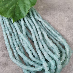 Könnte beinhalten: Eine Ansammlung mehrerer Stränge aus hellblauen, zylindrischen Perlen mit mattem Finish. Die Perlen haben eine leicht unregelmäßige, organische Form und sind aufgereiht. Ein einzelnes großes, grünes Blatt mit gezacktem Rand liegt auf den Perlen.